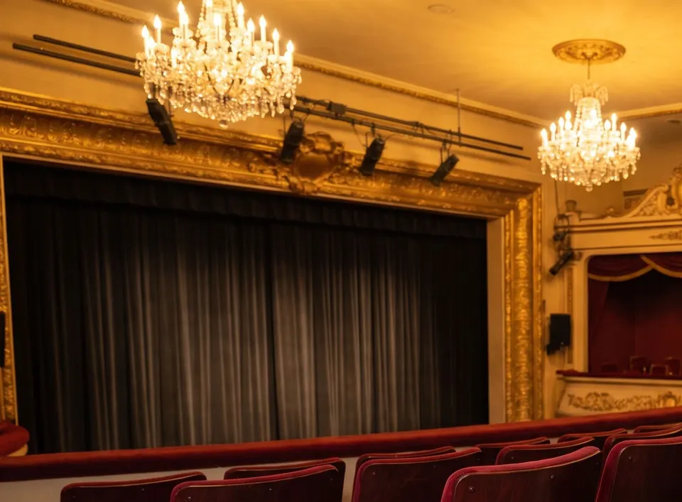 An empty small theatre before a show: a dark closed curtain in a gilded frame, crystal chandeliers and rows of red velvet seats.